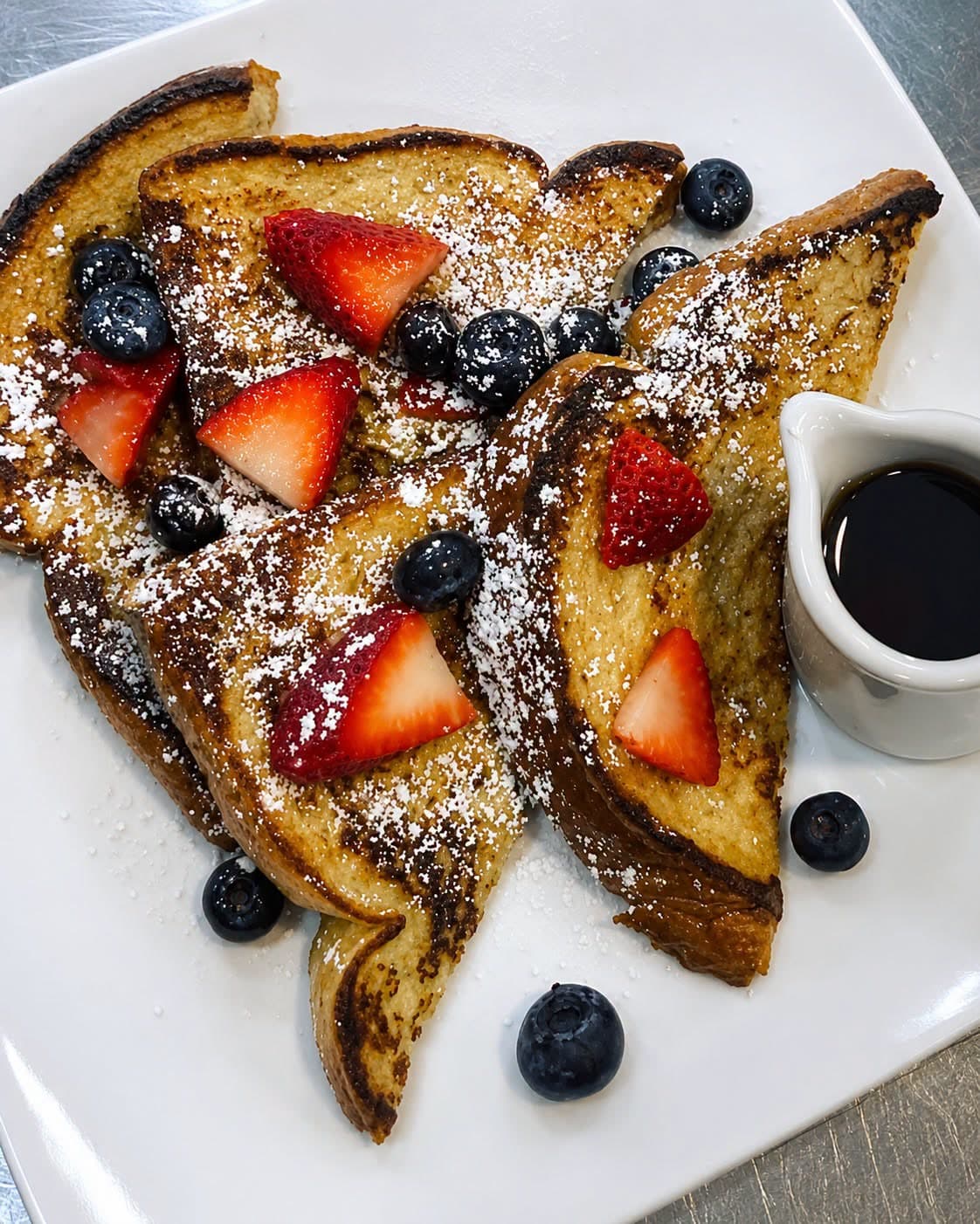 French toast triangles topped with sliced strawberries and blueberries, dusted with powdered sugar, with a pitcher of syrup