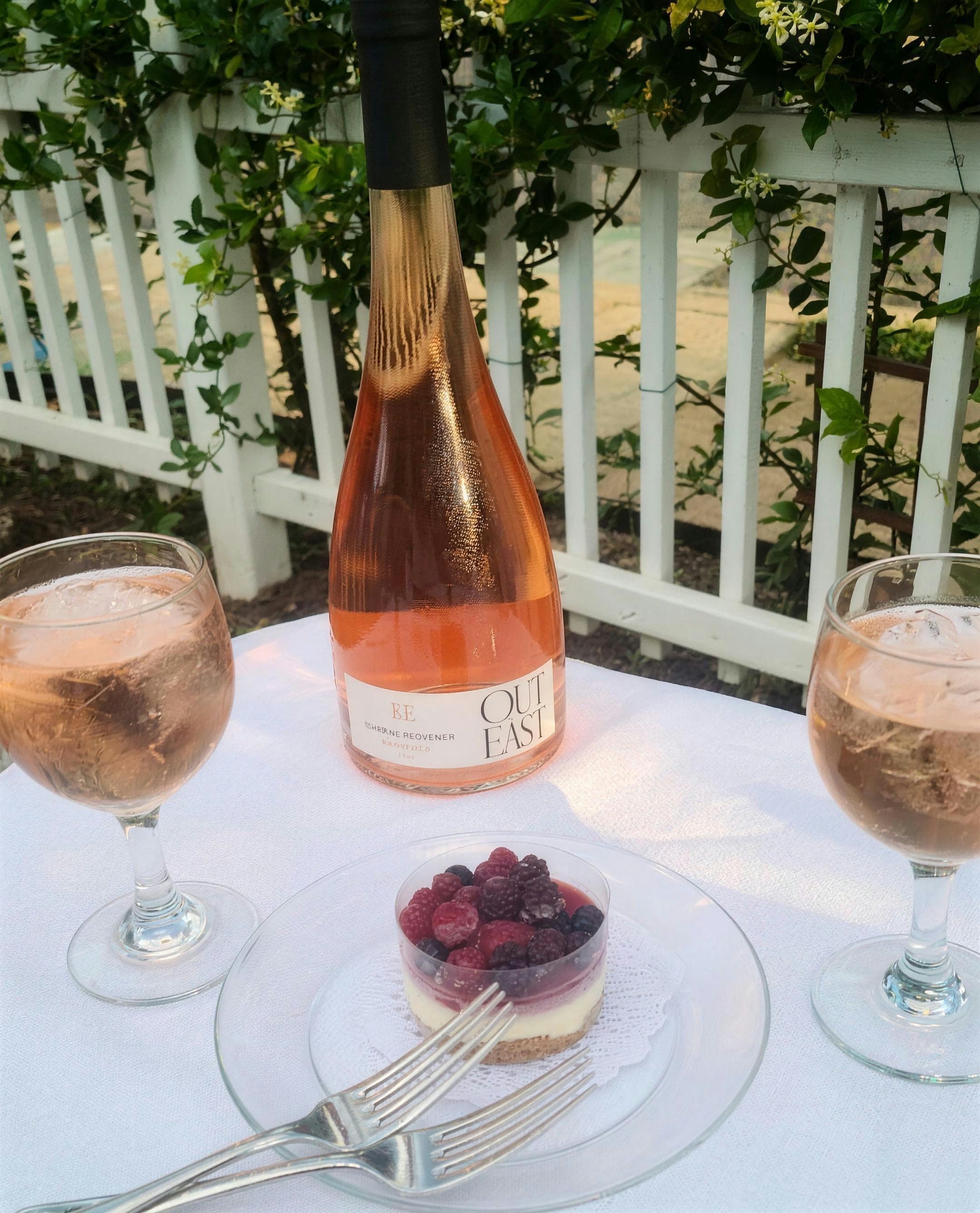 A bottle of Provence rosé and two filled glasses on a white-clothed patio table with a berry tart, against the white picket railing and jasmine of the terrace