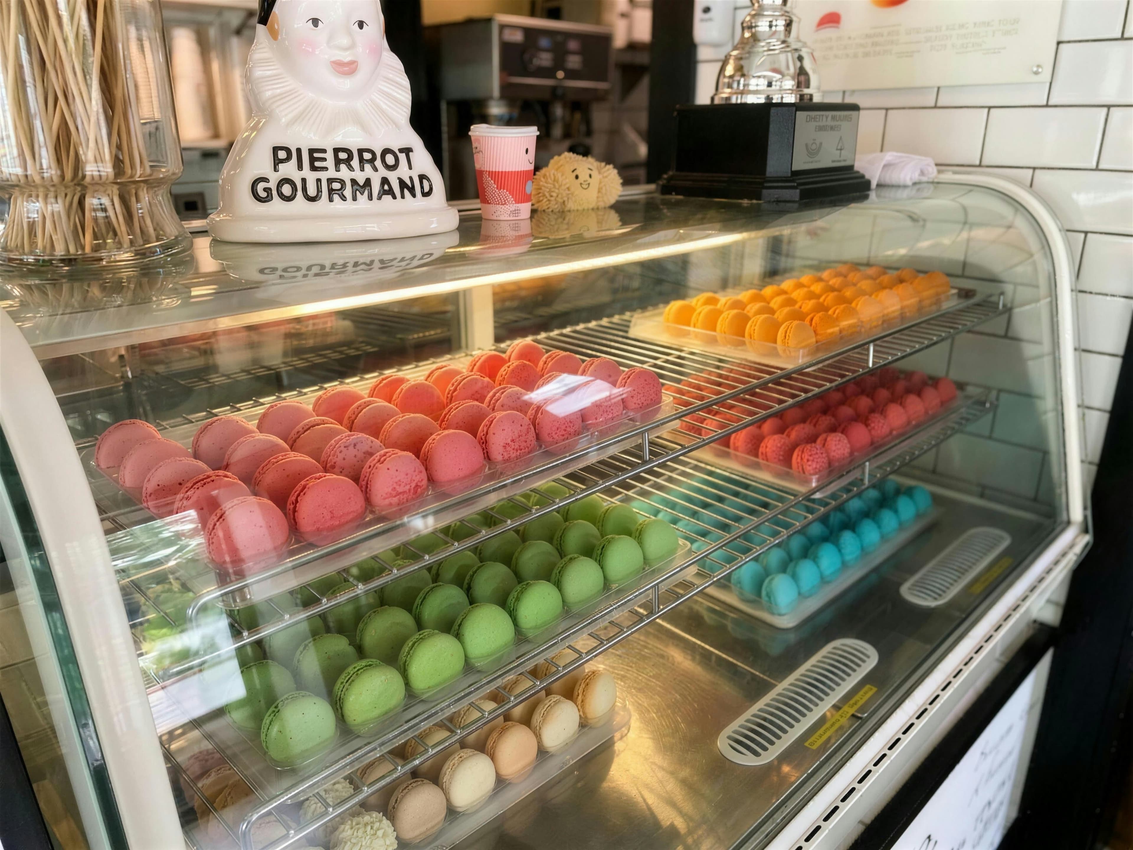 The macaron case — five colors on racks beside a Pierrot Gourmand bust and the Bienvenue Welcome counter